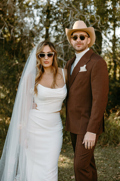 Bride and groom posing with sunglasses on with veil to the side and groom in cowboy hat
