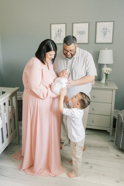 Mom in pink dress holding baby while dad and older brother look at newborn baby sister in their home