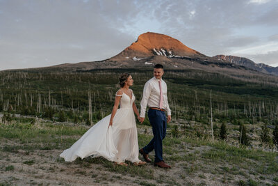 A couple holds hands and walks with an incredible mountain with orange alpenglow behind them during their elopement in Montana.