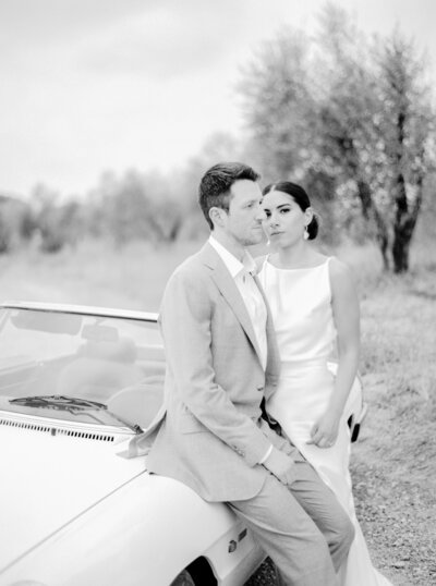black and white film photograph of bride and groom in Tuscany among olive trees and leaning on vintage white car photographed by Italy wedding photographer