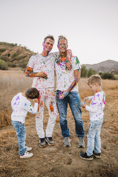 Gay couple and twin boys smile at camera covered in paint from paint fight session