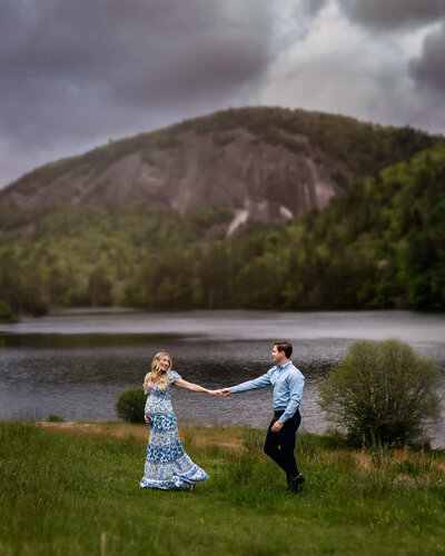 An expecting young woman in a beautiful long dress pulls her husband along a meadow with tall grass with mountains in the distance