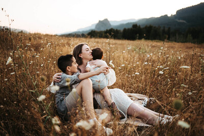 Mom with brown hair is sitting on  a wild flower field and hugging her toddler and pre teen age boy