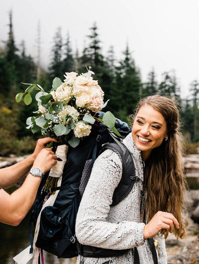 a bride gets help putting her wedding bouquet on her hiking pack for her pnw adventure elopement