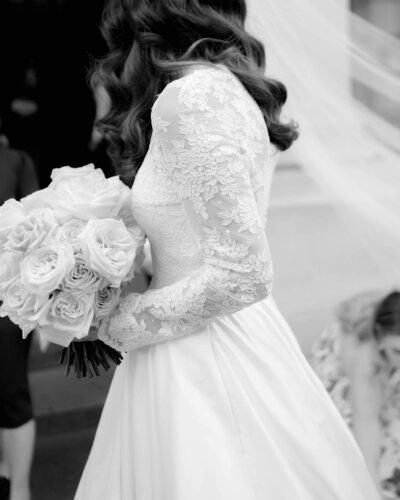 bride’s side profile showing her lace wedding dress sleeves as she holds a white floral bouquet and walks at her luxury wedding