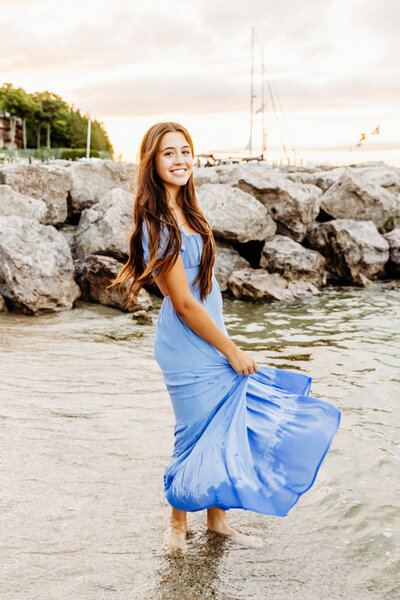 beautiful senior in a light blue dress standing in the water and spinning in her dress in Lake Michigan