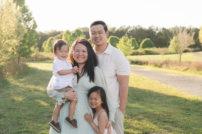 Korean family of four, mom holding 18 month old boy in right arm with left arm around five year old daughter and dad behind her left shoulder all standing in a park, smiling at the viewer during sunset
