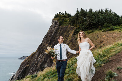A blond bride wearing a strapless white gown holds it up while walking through the muddy trail in her hiking boots while her groom in glasses holds her waist and carries a dried floral bouquet while looking at his bride as they hike down the Elk Flats Trail near Manzanita Oregon for their adventure elopement on the coast. | Erica Swantek Photography