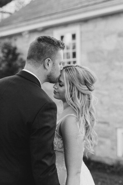 A black and white portrait of a groom kissing his bride's forehead