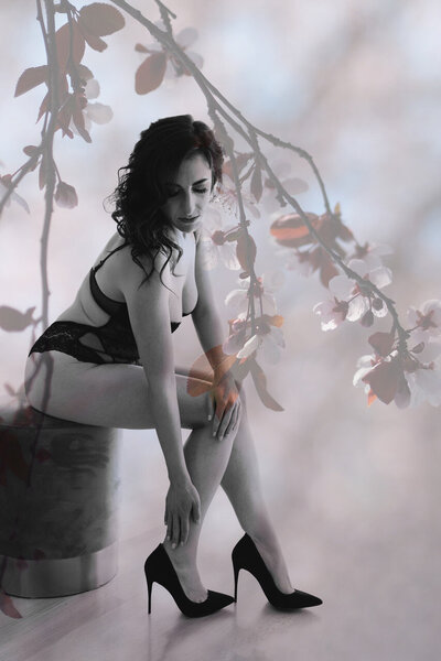 A black and white boudoir image from a San Fransisco boudoir photographer showing a woman in a side view as she sits on a stool, leaning over and running her hand down her leg, with one leg slightly forward. The artistic plant overlay adds depth and a unique artistic touch to the image.