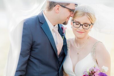 groom in dark blue suit whispers in bride's ear while bride wearing glasses , tiara and a veil smiles