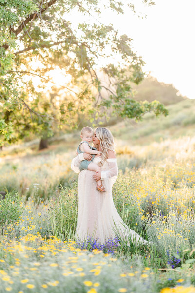 A family walks in a field of poppy flowers, the mother dressed in a long white gown holding her toddler sons hand photographed by Bay area Family photographer, Light Livin Photography.