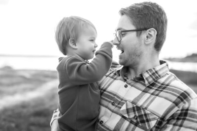 Black and white photo of a father and son. The dad is wearing reading black glasses and shares a playful moment with his baby boy who curiously plays with the glasses.