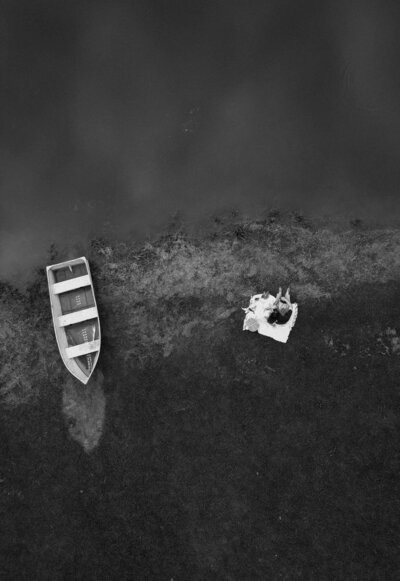 An overhead shot captures the intimate scene of a couple sitting on a blanket by the riverbank, with a boat nearby