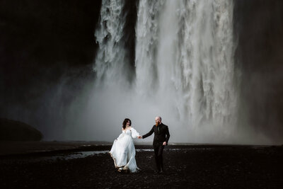 Married couple is holding hands and running in front of the Skogafoss waterfall in Iceland