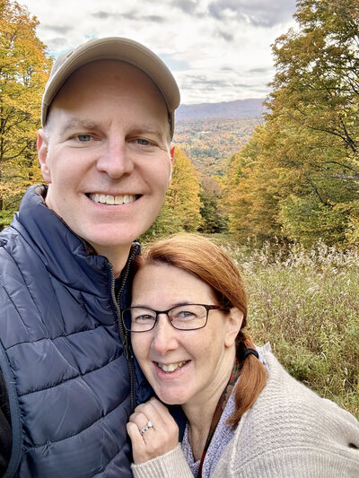 Carolyn Ann Ryan and her husband snuggle for a photo together while on vacation in Vermonth with fall foliage and mountain views in the background