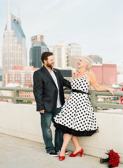 Bride in a 1950s white polkadotted pinup dress and a groom in jeans and a black silk suit jacket post on the pedestrian bridge with the Nashville skyline