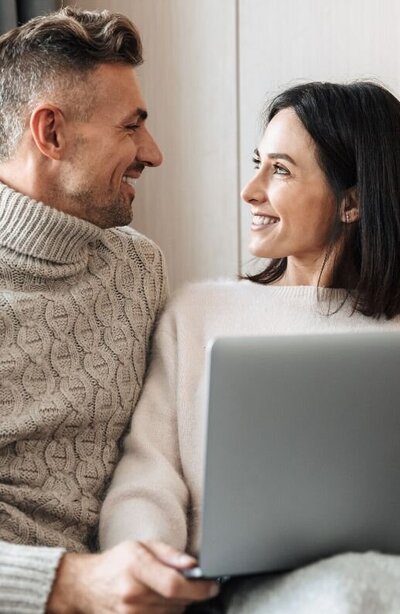 A couple smile at one another while holding a laptop. This could represent having an online couples therapy session from the comfort of their home. We offer online relationship therapy in Florida, online couples therapy, online couples counseling, and other services.