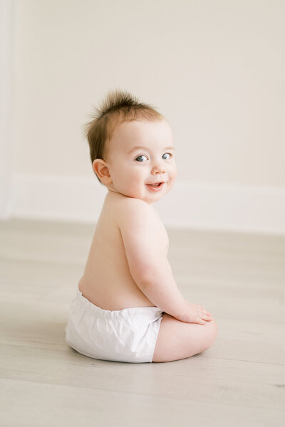 Baby boy sits up wearing a white diaper cover and looks at camera over his shoulder during studio photo session with Worth Capturing Photography.