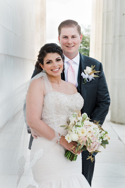 Photo of wedding couple standing in a walkway at the Lincoln Memorial surrounded by marble columns.The groom is standing behind the bride with her veil draped over her shoulder and arm while holding a beautiful bouquet of light colored roses.