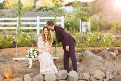 Groom kisses his Bride as she sits on a wood swing at the Forever & Always Farm wedding venue