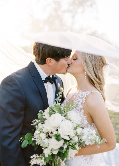 Groom in navy tux and bowtie kisses his blonde bride with lace high neck bridal gown. She is holding a green and ivory bouquet in front of them and her sheer veil is flowing in the wind above their heads at the gorgeous Metter, Ga wedding venue, Dutch Ford Farms.