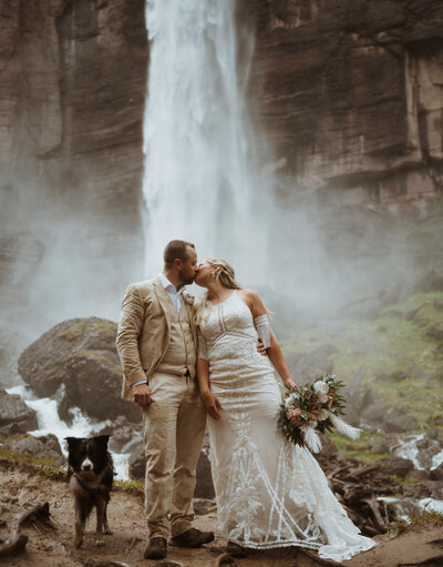 bride and groom are at the base of a waterfall in telluride. they are with their dog, holding hands and kissing. the bride has her bouquet, and both are getting wet from the mist at the waterfall.