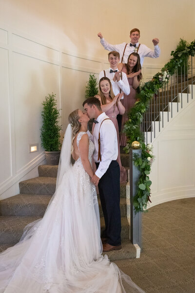 Bride and groom kissing at bottom of stairs with wedding party behind them
