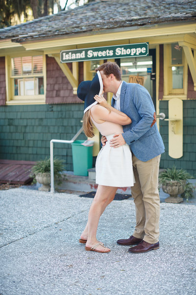 Bride Cups Floppy Hat around Face as Fiance Kisses her in Front of a Candy Shoppe at their Jekyll Island Engagement Photos