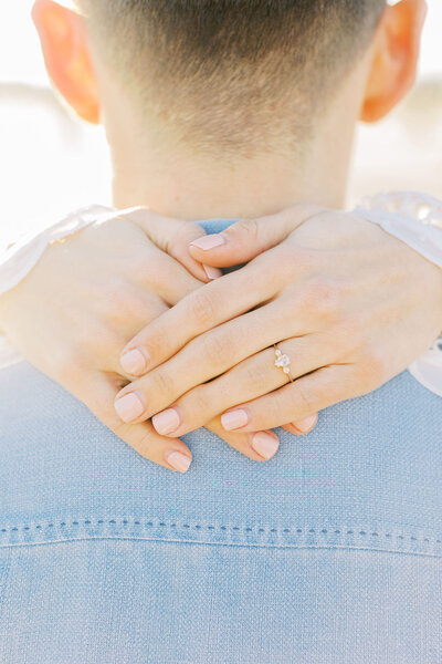 Close-up photo of the engagement ring on her hands wrapped around his neck as she hugs her fiance from behind.