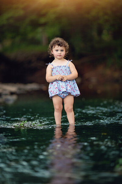 A toddler girl in a floral print bathing suit stands in a shallow flowing river