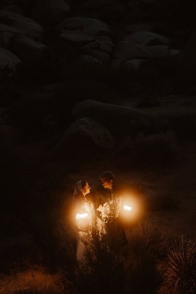 Two women hold hands and smile at each other during golden hour, standing on dark volcanic rock with desert plants around them in the Mojave Desert.