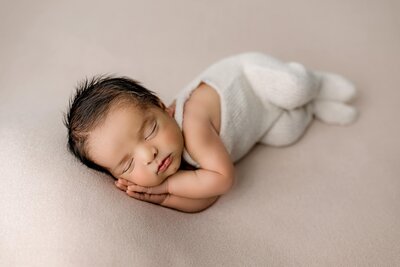 Baby boys dressed in cream overalls laying on his side with both hands underneath his cheek on a cream background in their Studio newborn session