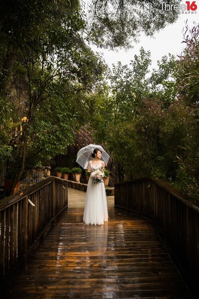 Bride stands on the deck at Calamigos Ranch under an umbrella as the rain drops down