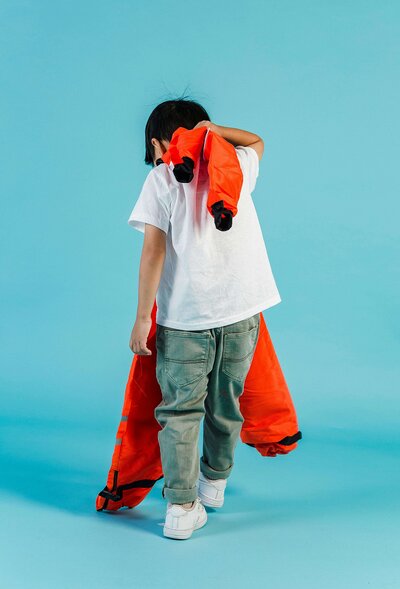 A young child with their back turned, wearing a white t-shirt and jeans, is draping a bright orange jacket over their shoulder against a blue background.