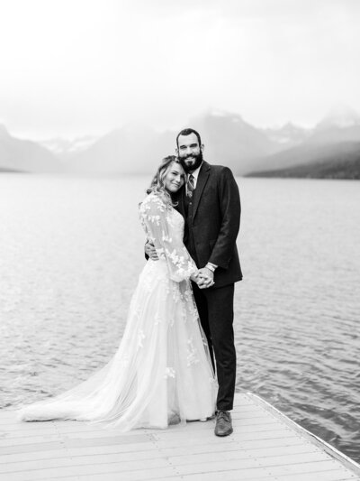 Black and white photo of the bride and groom posing by the water with mountains behind them at their Glacier National Park wedding.