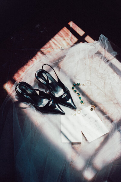 Wedding details photographed in soft window light, including black heels, a veil, and emerald jewelry.
