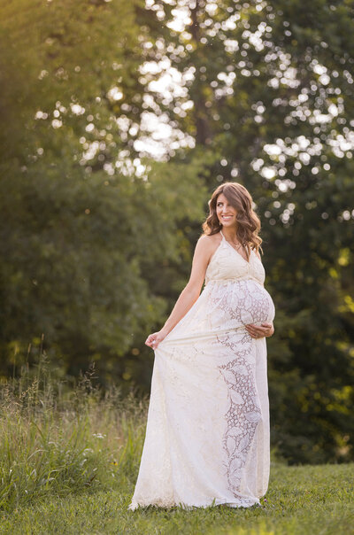 Maternity photo of expectant mother in cream lace gown with hand under belly with backlight on her brown hair