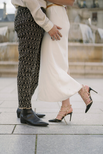 the bottom half of a man and woman in dress clothing facing one another in front of a fountain in paris with the woman kicking up one foot in her valentino heels
