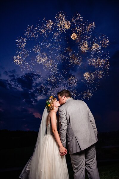 Bride wearing a flower crown holding her husbands hand and they are kissing while fireworks go off behind them