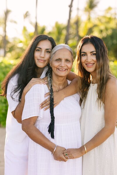 Three generations of women hold hands posing for an image on the beach.