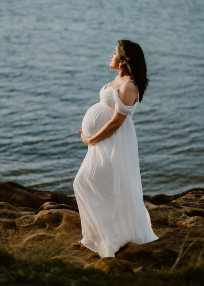 First time mum to me wearing a white long dress on a cliff gazing out towards the blue ocean.