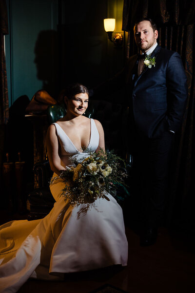 Dramatic portrait of a bride seated in an antique chair, with the groom standing beside her, both in sophisticated wedding attire, in the Lord Thompson Manor