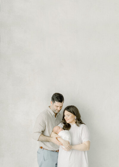 A mother standing by hand painted backdrop in a Dallas photography studio holding her newborn baby as her husband stands behind her holding them both.