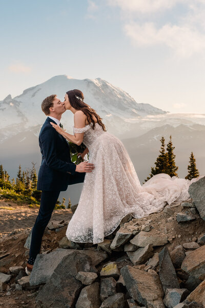 after learning how to elope in Washington State, a couple in their wedding attire kisses while standing on boulders with Mount Rainier in the background