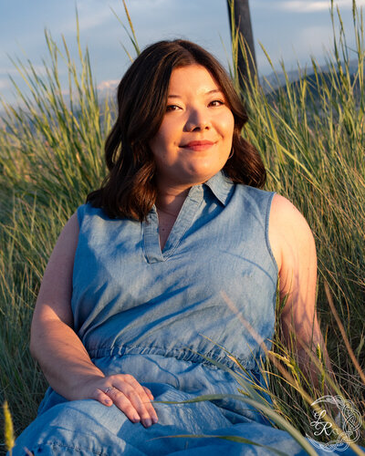 Photo of Ashlyn sitting in the grass in a blue denim dress with her hand in her lap