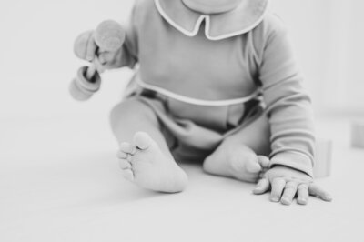 Black and white close up of baby's foot and hand holding wooden rattle during photo session at Worth Capturing studio