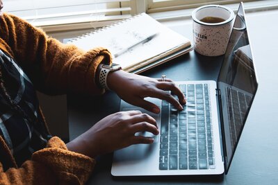 Hands of a dark-skinned woman at a laptop keyboard, with a notebook and coffee nearby. Photo by Daniel Thomas via Unsplash.
