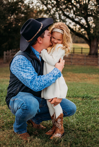 A League City dad tickles and kisses the cheek of his toddler daughter who wears a white dress and cowboy boots.