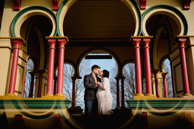 A pregnant person and their partner leaning in for a kiss while standing under a colorful gazebo.